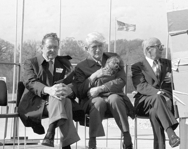 Yes, that is indeed a dog Ramsey is holding in his coat on the fiercely windy Saturday, May 11, 1974 when the National Accelerator Laboratory was renamed Fermi National Accelerator Laboratory. The dog belonged to Dixy Lee Ray, chair of the U.S. Atomic Energy Commision. 'I used to jokingly point out that this was the primary responsibility of the president of the URA,' Ramsey said. 'The auditorium wasn't finished yet, and we asked the Italian priest from Chicago, who was advocating Fermi renaming, figuring he might have a direct line. He said it would stop raining, but we didn't ask him the right question. The wind came up vastly greater. We were worried about the temporary podium blowing over. It came time for Dixy Lee Ray to talk, and the question was what to do with her dog, who went everywhere with her, and I volunteered to hold it on my lap.'